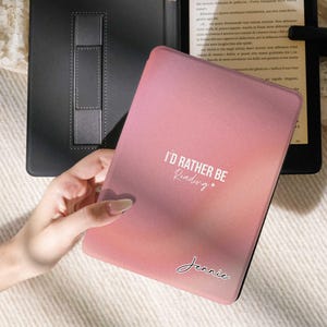 Peut inclure: Une housse de liseuse rose et pêche en dégradé avec le texte "I'D RATHER BE Reading" et le nom "Jennie" en écriture cursive. L'étui est ouvert, révélant un lecteur électronique noir. Un support de liseuse noir est visible en arrière-plan.