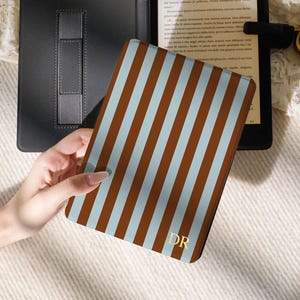 May include: A protective e-reader case featuring a vertical striped pattern in light blue and brown. The front cover displays the gold-coloured initials DR in the bottom right corner. The case is shown open, revealing a black interior frame.