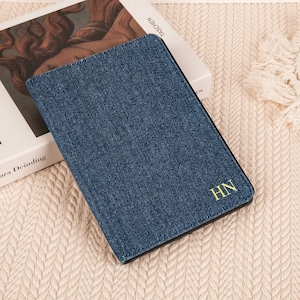 Peut inclure: Un porte-passeport ou une couverture de carnet rectangulaire avec un extérieur en tissu denim bleu foncé. Le coin inférieur droit présente les initiales HN brodées en fil doré avec une police serif. L'objet a une bordure noire et un profil fin.