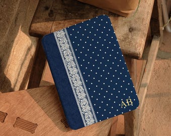 Pode incluir: Uma capa retangular para leitor de e-books em azul-marinho com um padrão de bolinhas brancas, uma faixa vertical de renda floral branca no lado esquerdo e as letras do monograma AB em dourado impressas no canto inferior direito da capa.