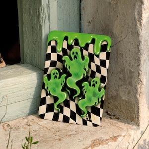 May include: A rectangular sign featuring a black and white checkered pattern background with three translucent green ghosts floating in the center. The top edge has a green dripping paint effect, and the letters AB are printed in the bottom right corner.