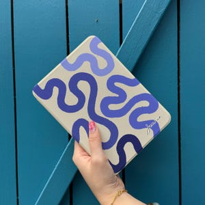 May include: A tablet case with a beige background and a repeating pattern of blue and purple wavy lines. The name "Jeanie" is written in cursive in the lower right corner. The case is held against a blue wooden background.