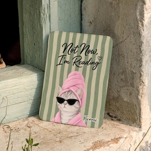 May include: A rectangular decorative sign with a cat wearing a pink towel wrap and sunglasses. The sign has the text "Not Now, I'm Reading" and the name "Jennie". The background has green and white vertical stripes.