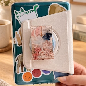 May include: A dark teal tablet case decorated with various stickers, including a cat, a candle, and smiley faces. A light beige magnetic cover is attached, featuring a rectangular sticker of a sword, flowers, and the text You could rattle the stars. To whatever end. You do not yield.