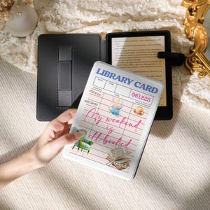 以下が含まれることがあります： 「My weekend is all booked」のテキストが入った「Library Card」デザインの白い電子書籍リーダーカバー。カバーはピンク色のグリッドの背景に、ティーカップ、アームチェア、本の水彩イラストが描かれています。背景には黒い電子書籍リーダーが見えます。