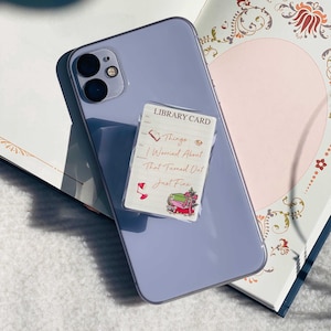 May include: A light purple smartphone with a clear case, featuring a library card-themed phone grip. The grip has the text "Things I Worried About That Turned Out Just Fine" with illustrations of books and a coffee cup.