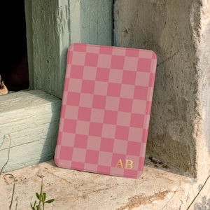 May include: A rectangular tablet or e-reader case featuring a pink and light pink checkered pattern. The bottom right corner of the case displays the gold-colored initials AB in a serif font. The case has rounded corners and a smooth finish.