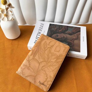 以下が含まれることがあります： 花柄の模様が施された茶色の革製のノートブックで、表紙には「Sydney」という名前が刻印されています。ノートブックは、茶色の表面に置かれ、タッシェン社の「ボッティチェリ」というタイトルの本の隣にあります。