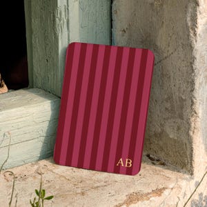 May include: A rectangular tablet case featuring a vertical striped pattern in alternating shades of dark red and burgundy. The bottom right corner of the case displays the gold-colored capital letters AB in a serif font, centered on the striped surface.