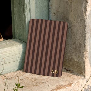 May include: A rectangular brown case with vertical stripes in varying shades of brown. The initials "AB" are in gold at the bottom right corner. The case is leaning against a weathered, light green wooden frame and a stone surface.