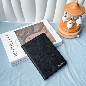 以下が含まれることがあります： 黒い革のジャーナルで、花柄のエンボス加工が施され、前面に白い文字で「アンドレア」と書いてあります。ジャーナルは薄い青色の表面に置かれ、バルバラ・ドムリング著の「ボッティチェリ」というタイトルの本の隣にあります。