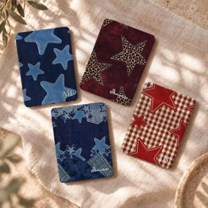 May include: Four rectangular passport holders featuring star patterns. One is dark blue with light blue stars and initials AB, one is maroon with leopard print stars, one is denim patchwork with stars, and one is red and white gingham with red stars.