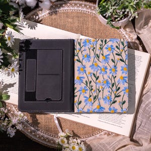 May include: Open black journal with a floral embroidered cover featuring blue and yellow daisies on a beige background. The journal is open, revealing a pocket and a pen holder. The journal is lying on top of an open book.