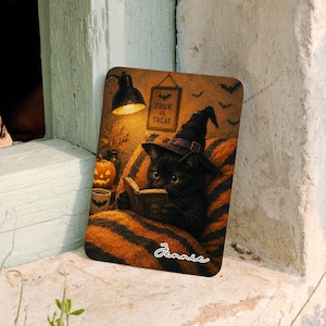 May include: A rectangular sign featuring a black cat wearing a witch hat and reading a book while sitting on an orange and black striped blanket. A lit lamp, a jack-o'-lantern, and a sign reading Trick or Treat are visible. The name Jennie is printed.