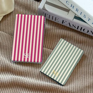 May include: Two rectangular passport holders featuring vertical stripes. One holder has magenta and white stripes with the monogram SB in white. The other holder has sage green and white stripes with the monogram NE in gold. Both items are flat.
