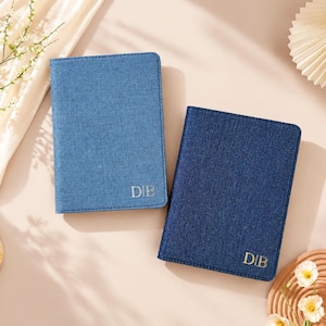 May include: Two rectangular passport holders with a denim-like texture. One is light blue and the other is dark blue. Both feature the gold-colored embossed initials D|B in the bottom right corner. The covers have stitched edges.