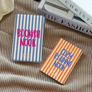 May include: Two rectangular e-reader cases with vertical stripes. The left case is blue and white with the pink text "BOOKISH NOOK" and the gold initials "NE". The right case is orange and white with blue text "COZY READING NOOK" and "SB".