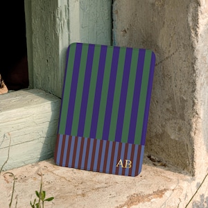 Pode incluir: Uma capa retangular para tablet com um padrão de riscas verticais em roxo escuro e verde floresta na parte superior, e um padrão de riscas verticais mais estreitas em azul escuro e bordeaux na parte inferior com as letras douradas AB.