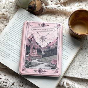 Puede incluir: Una funda para lector electrónico de color rosa claro con una ilustración detallada de un castillo, montañas y una estrella. El texto READ BENEATH THE STARS está impreso en la parte superior. La funda tiene motivos de rosas y bordes.