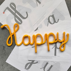 May include: A yellow knitted word "Happy" is displayed on top of a set of instructions for creating lettering and figures. The instructions are printed on white paper and include diagrams and arrows showing the direction of the yarn.