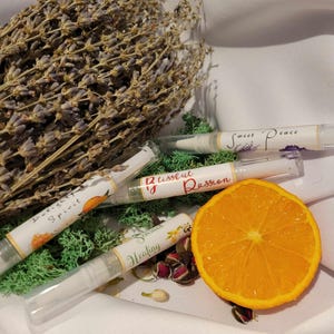 May include: Four clear glass roller bottles with labels. The labels have different colors and text. The text on the labels reads "Blissful Passion", "Sweet Peace", "Sensual Healing", and "Light Spirit". The bottles are on a white surface with a green moss, lavender flowers, and a slice of orange.