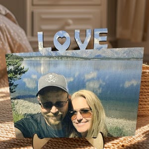 May include: A wooden photo frame with a couple's photo and the word "LOVE" with a heart cutout. The photo shows a couple in front of a lake and mountains.