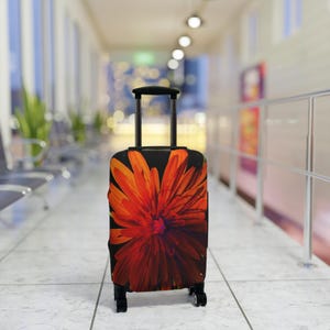 Op de afbeelding: Een zwarte rolkoffer met een levendig oranje en rood bloemenpatroon. De koffer heeft een telescopische handgreep en vier wielen. Het bloemenpatroon is een close-up van een bloem.