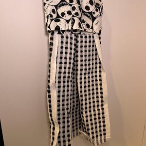 K&ouml;nnte beinhalten: Ein schwarz-wei&szlig;es K&uuml;chentuch mit einem Jack-Skellington-Sch&auml;delmuster oben. Das Handtuch hat ein kariertes Muster mit wei&szlig;em Rand und ist an einer Wand befestigt.