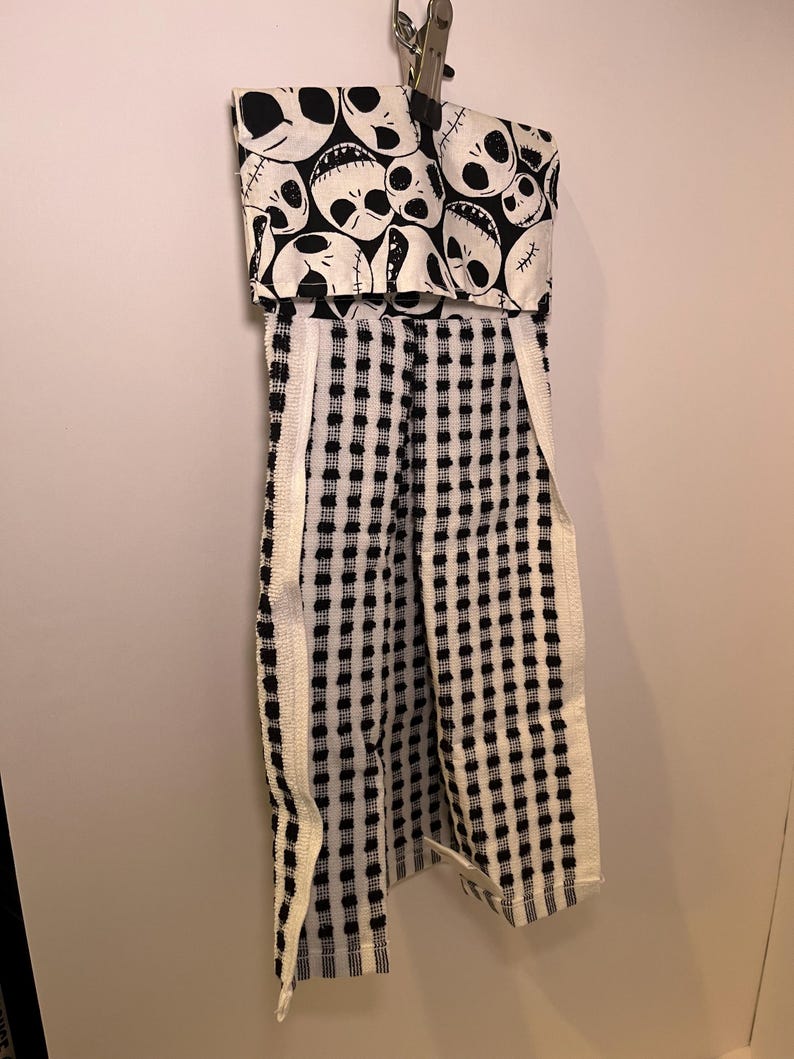 K&ouml;nnte beinhalten: Ein schwarz-wei&szlig;es K&uuml;chentuch mit einem Jack-Skellington-Sch&auml;delmuster oben. Das Handtuch hat ein kariertes Muster mit wei&szlig;em Rand und ist an einer Wand befestigt.