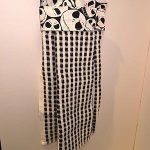 K&ouml;nnte beinhalten: Ein h&auml;ngendes K&uuml;chentuch mit einem schwarz-wei&szlig;en Karomuster. Der obere Teil hat ein umgefaltetes Design mit einem sich wiederholenden Muster von Jack Skellingtons Gesicht aus The Nightmare Before Christmas. Das Handtuch h&auml;ngt an einer Metallklammer.