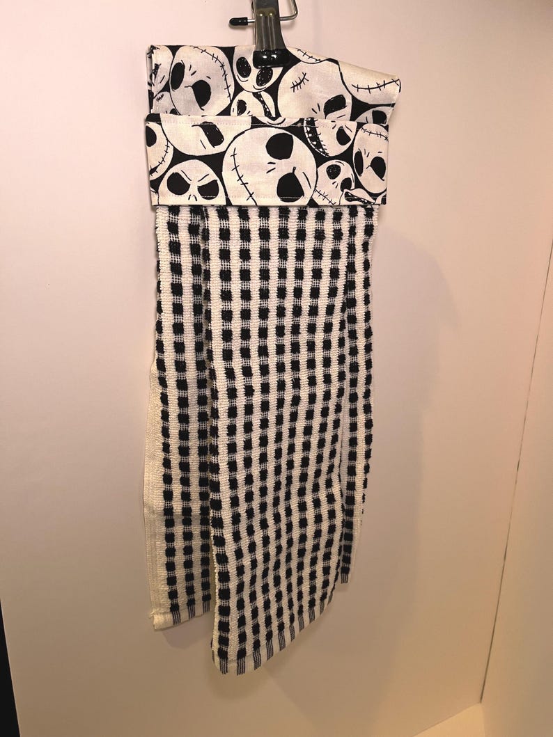 K&ouml;nnte beinhalten: Ein h&auml;ngendes K&uuml;chentuch mit einem schwarz-wei&szlig;en Karomuster. Der obere Teil hat ein umgefaltetes Design mit einem sich wiederholenden Muster von Jack Skellingtons Gesicht aus The Nightmare Before Christmas. Das Handtuch h&auml;ngt an einer Metallklammer.