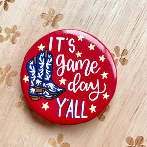 May include: Red button with a white design that says "It's game day y'all" with a pair of blue and white cowboy boots. There are yellow stars around the text and boots.