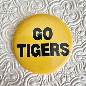 May include: A round, yellow button with the words "GO TIGERS" in bold, black letters. The button is set against a textured, white background.