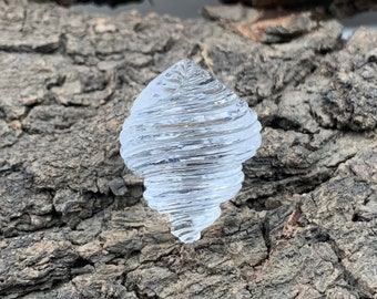 Puede incluir: Un objeto de vidrio transparente e incoloro con forma de concha marina en espiral. La superficie presenta crestas horizontales en relieve que envuelven la forma cónica, creando una apariencia texturizada y acanalada con un acabado brillante.