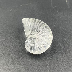 Puede incluir: Escultura de concha de nautilus de vidrio transparente. La concha presenta una textura acanalada detallada, mostrando un dise&ntilde;o en espiral. La escultura est&aacute; sobre una superficie oscura, resaltando su forma transparente.