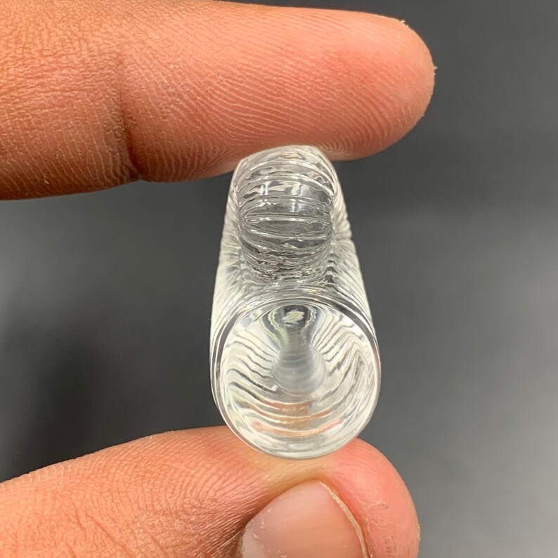 Puede incluir: Un objeto de vidrio transparente y cil&iacute;ndrico con una superficie texturizada. El objeto tiene una parte superior redondeada y una base acampanada. La textura parece ser una serie de l&iacute;neas curvas. El objeto se sostiene entre dos dedos.