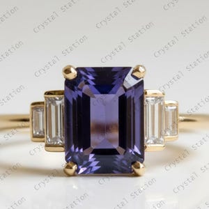 Può includere: Un anello d'oro con una grande gemma rettangolare viola scuro come elemento centrale. Ai lati della pietra principale ci sono gemme chiare rettangolari più piccole. L'anello ha un design classico ed elegante.