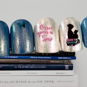 May include: Five fingernails painted with a light blue glitter polish. One nail has a small blue butterfly sticker, one nail has the words "Once upon a Time" written in pink, one nail has a silhouette of a person reading a book, and two nails are plain.