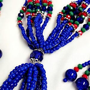 May include: A blue beaded necklace with a tassel and silver accents. The necklace features blue, green, and red beads.