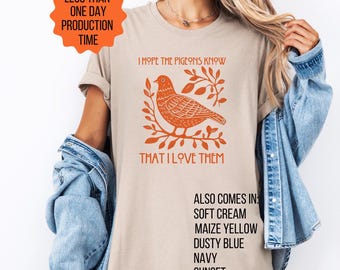 May include: Beige t-shirt with an orange graphic of a bird on a branch, accompanied by the text "I HOPE THE PIGEONS KNOW THAT I LOVE THEM." Additional color options are listed below the graphic.