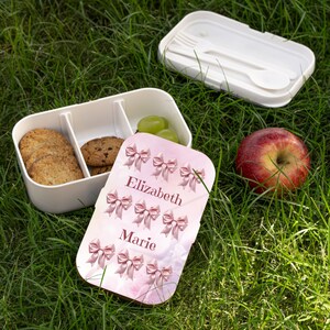 May include: A white lunchbox with a pink and white lid. The lid has a pink bow pattern and the names "Elizabeth" and "Marie" written on it. The lunchbox is open and contains cookies and grapes.