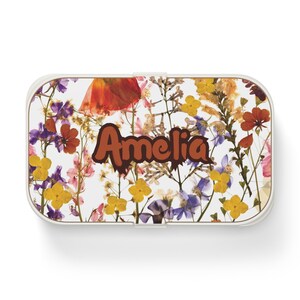 May include: A white lunchbox with a floral design featuring pressed flowers in shades of pink, purple, yellow, orange, and brown. The lunchbox has the name "Amelia" written in brown letters.