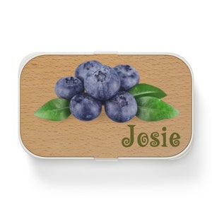 May include: A wooden lunchbox with a white border and a design of blueberries and green leaves. The name "Josie" is written in green cursive on the front.