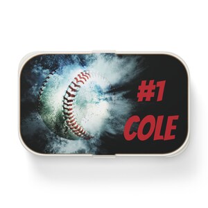 May include: A white and black rectangular lunchbox featuring a baseball design. The baseball is white with red stitching, set against a smoky blue background. The text "#1 COLE" is in red.