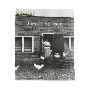 May include: A black and white photo of a woman standing in front of a brick building with the words "Local Egg Dealer" above the door. She is holding a basket of eggs and there are chickens in the yard.