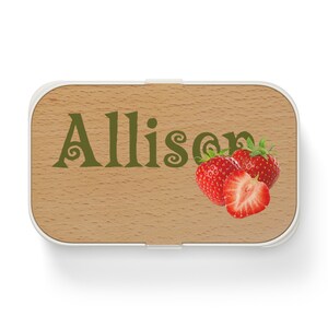 May include: A wooden lunchbox with a white lid. The lunchbox has the name "Allison" written on it in green letters. There are three red strawberries on the lunchbox.