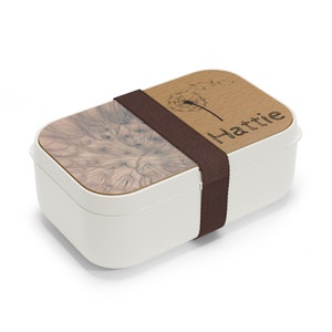 May include: White lunchbox with a brown strap and a wooden lid. The lid has a dandelion design and the name "Hattie" engraved on it.