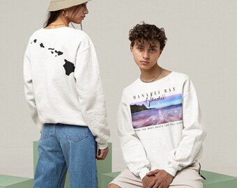 May include: Two white sweatshirts with different designs. One sweatshirt has a black outline of the Hawaiian islands on the back. The other sweatshirt has a colorful image of a beach scene with the text "Hanalei Bay Hawaii" and "Where the moon meets the sea, Kauai".