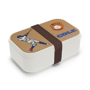 May include: White lunchbox with a brown wooden lid featuring a baseball player graphic and the text "COLE". The lid also has a baseball and glove graphic. The lunchbox has a brown strap.