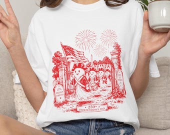 May include: White t-shirt featuring a red graphic commemorating the 250th Independence Day. The design includes a parade of figures, an American flag, fireworks, and the text "1776 Freedom Forever".